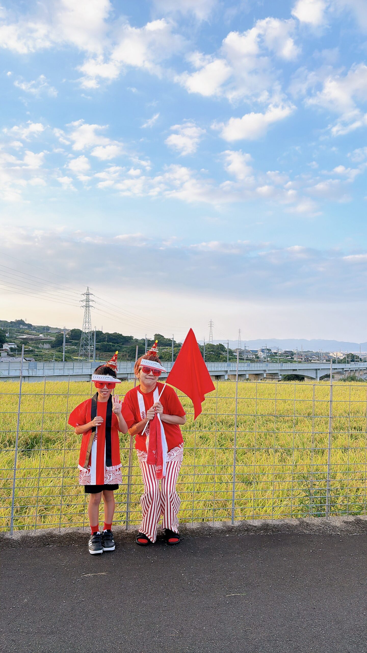 田んぼと線路を背景に、赤い服と旗で西九州新幹線かもめを待つ子どもと大人