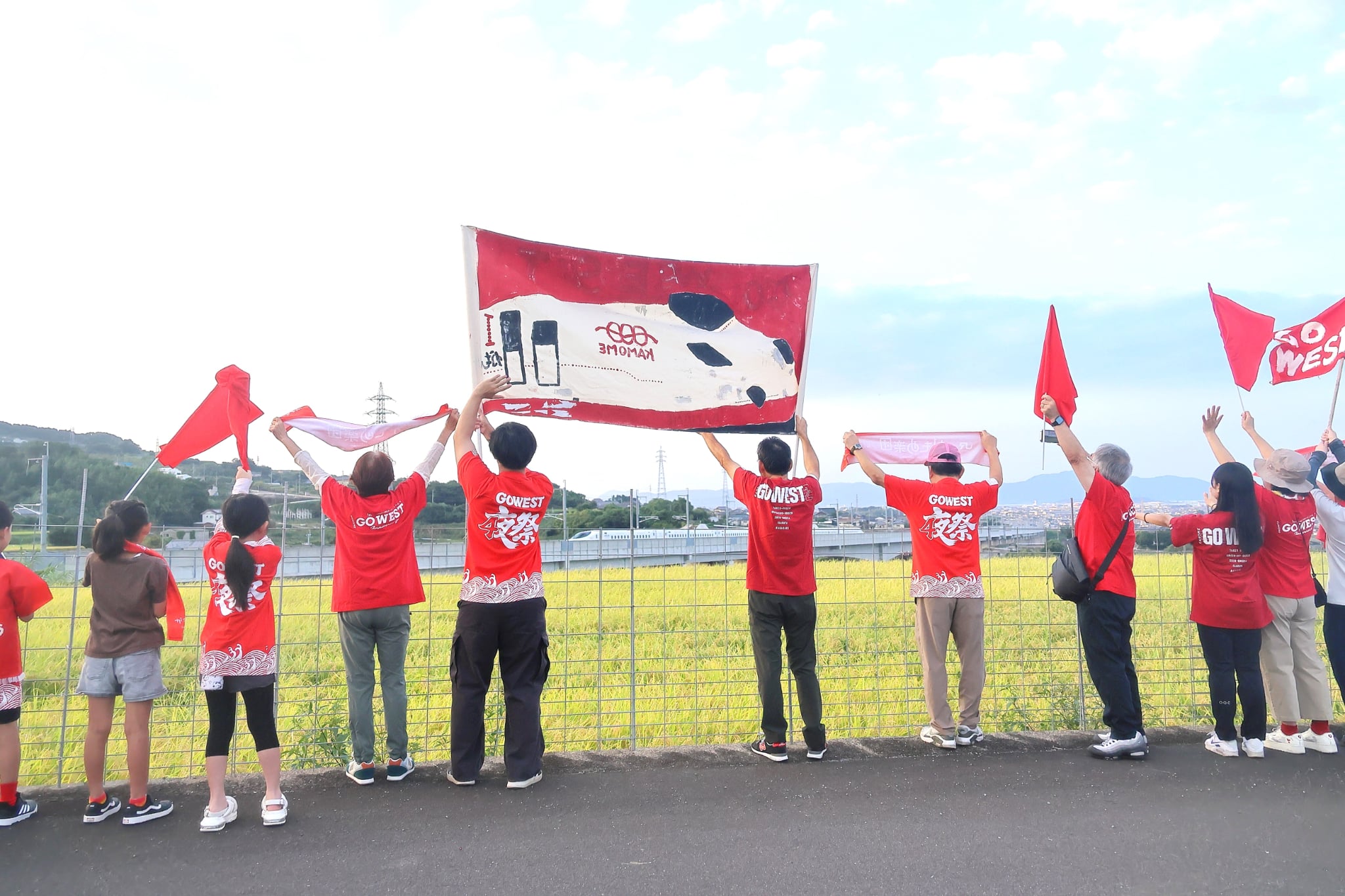 田んぼと線路に向かって旗を掲げる子どもと大人たち